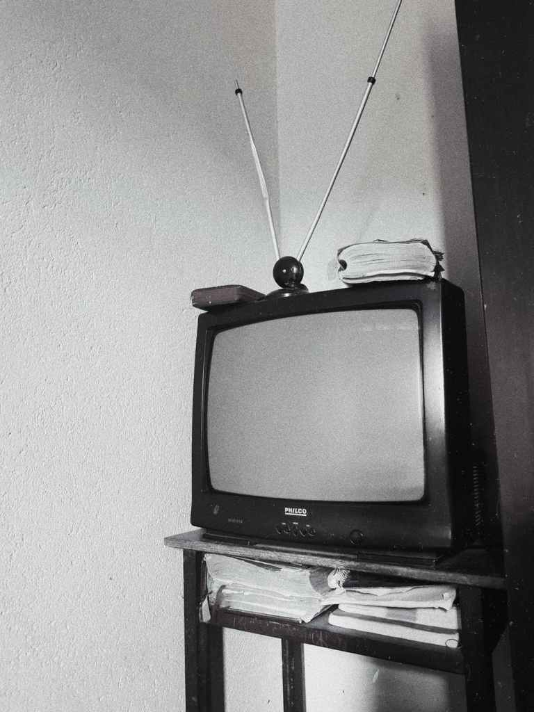 Uma televisão antiga desligada sobre uma pequena mesa, com livros muito gastos sob ela. Acima da TV há uma antena interna, mais um livro muito usado e o que parece ser uma carteira. Parece remeter à uma casa de família de classe média baixa nos anos 80.